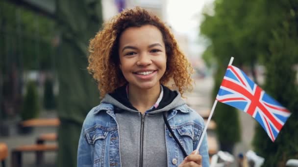 Zeitlupenporträt einer jungen und hübschen afrikanisch-amerikanischen Dame mit britischer Flagge und lächelndem Blick in die Kamera an einem windigen Tag. Touristisches und attraktives Konzept. — Stockvideo