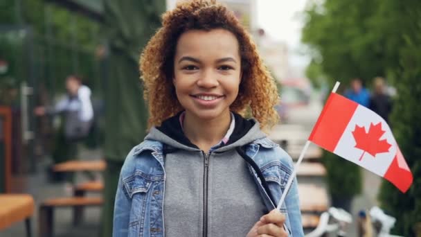 Afro-Amerikan kız gezgin holding Kanada bayrağı gülümseyen ve kamera açık havada seyir Slowmotion portre. Mutlu turist ve ziyaret yabancı coutries kavramı. — Stok video