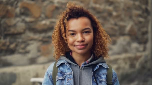 Cerrar retrato en cámara lenta de la niña afroamericana riendo con el pelo rizado claro en chaqueta de mezclilla mirando a la cámara y posando. Jóvenes felices y concepto de estilo de vida moderno . — Vídeos de Stock