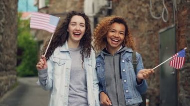 İki güzel kız Amerikan bayrakları sallayarak ve kameraya bakarak gülen casual giyim Slowmotion portresi. Dostluk, turizm ve mutlu insanlar kavramı.