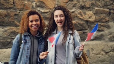 Slowmotion Fransız bayrakları sallayarak ve kamera ayakta brickwall karşı bakarak iki gülen kadınlar öğrenci portresi. Dostluk, turizm ve mutlu insanlar kavramı.