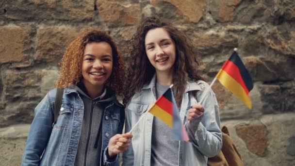 Portrait ralenti de deux amies souriantes agitant des drapeaux allemands et regardant la caméra debout contre un mur de pierre. Amitié, tourisme et personnes heureuses concept . — Video