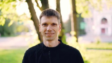 Ağır çekim yakın çekim portre neşeli genç adam gülümseyerek ve kamera açık havada arıyorum. Güzel doğa, sıcak güneşli gün, ağaçlar ve arka planda çimen.