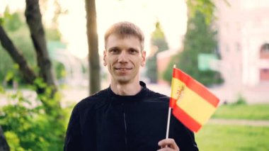 İspanyol bayrağı sallayarak ve ayakta ağaçlar, yeşil çim ve güzel binalar arkasında güzel parkta gülümseyen mutlu turist genç adam portresi ağır çekim.