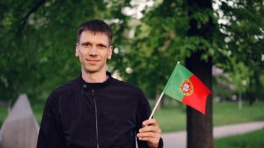 Portekiz bayrağı sallayarak ve Şehir Parkı yüksek ağaçları ve güzel çimenler ile ayakta gülümseyen neşeli genç adam gurur vatansever ağır çekim portre. İnsanlar ve ülkeler kavramı.