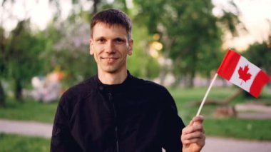 Yavaş hareket Kanada bayrağı tutarak ve kameraya bakarak ve gülümseyen sallayarak mutlu yakışıklı adam portresi. Arka planda, kişi odaklı yeşil şehir parkı olduğunu.