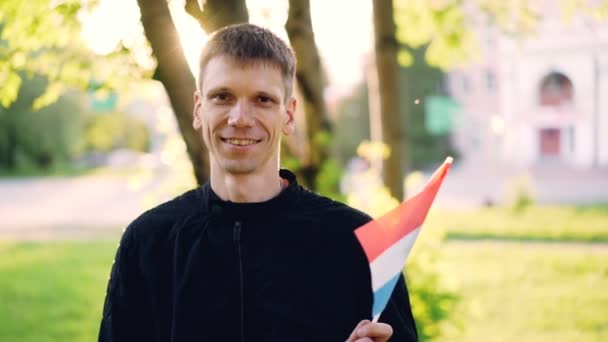 Retrato en cámara lenta del joven ondeando la bandera de los Países Bajos, sonriendo y mirando a la cámara con árboles, césped verde y hermoso edificio en el fondo . — Vídeo de stock