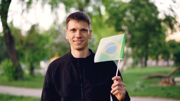 Slow motion portrait of glad traveller young man holding Brazilian flag and smiling looking at camera. National symbol, proud people, summertime and nature concept. — Stock Video