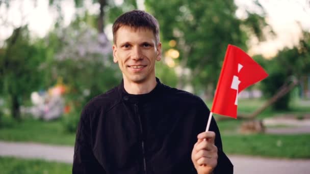 Retrato en cámara lenta de un joven viajero alegre sosteniendo la bandera nacional suiza, mirando a la cámara y sonriendo. Patriotismo, viajes por el mundo, gente feliz y concepto de naturaleza . — Vídeos de Stock