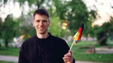 Ağır çekim portre resmi bayrak sallayarak ve kamera güzel ağaçlar ve çimenler büyük modern park dururken bakarak neşeli Alman Fan.