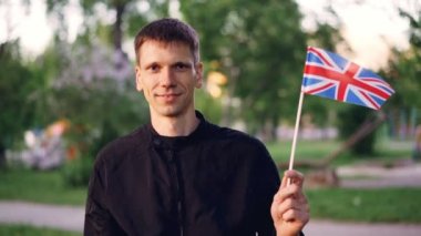 Genç adam gurur vatandaş holding İngiliz bayrağı önünde duran streen ile arka planda güzel park ve kameraya bakarak gülen, ağır çekim portre.