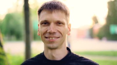 Ağır çekim yakın çekim portre kısa siyah saçlı ve kahverengi gözleri açık havada duran ile gülümseyen genç adamın kişi üzerinde odaklanmak arka planda, doğa ile kameraya bakıyor.