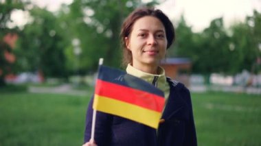 Ağır çekim portre resmi Alman bayrağı sallayarak ve kamera güzel ağaçlar ve çimleri güzel yeşil park dururken bakarak neşeli genç kadın.