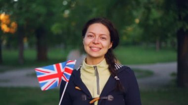 Ağır çekim Birleşik Krallık Ulusal bayrak sallayarak rahat kıyafetler içinde gülümseyen ve arka planda park ile kameraya bakarak neşeli genç bayan portresi.