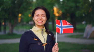 Yavaş hareket Ulusal Park ağaç ve yeşil çimenler ayakta ve gülümseyen Norveç bayrağı sallayarak oldukça Norveçli kadın portresi. İnsanlar ve ülkeler kavramı.