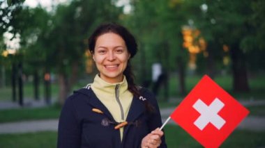 Ağır çekim gülümseyen ve kameraya bakarak İsviçre ' nin bayrak sallayarak neşeli İsviçre kız portresi. Yüksek ağaçları ve yeşil çimenler güzel park arka planda olduğunu.