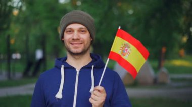 Ağır çekim portre sakallı genç adam İspanyol bayrağı sallayarak ve mutlu gülümseme konumu güzel yeşil şehir parkı olan kameraya bakıyor. İnsanlar ve Vatandaşlık kavramı.