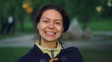 Ağır çekim portre çekici genç kadın dışarıda parkta duran ve kameraya bakarak gülen rahat kıyafetler giymiş. İnsan, Doğa ve sağlıklı yaşam konsepti.