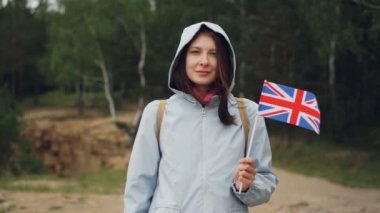 Gülümseyen ve kameraya bakarak Büyük Britanya bayrağı tutarak çekici İngiliz ağır çekim portre. Arka planda, rüzgarlı bahar günü yeşil ağaçlar ile güzel orman.