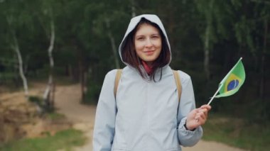 Ağır çekim portre gülümseyen ve kameraya bakarak rüzgarda uçan Brezilya bayrağı sallayarak güzel genç bayan memnun turist. Ülkeler ve insanlar kavramı.
