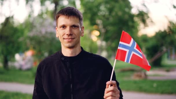 Retrato en cámara lenta de un estudiante noruego feliz sosteniendo la bandera nacional oficial, mirando a la cámara y sonriendo. Hermoso parque verde está en el fondo . — Vídeo de stock