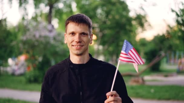 Retrato em câmera lenta de cara atraente em roupas casuais acenando bandeira americana, sorrindo e olhando para a câmera. Pessoas felizes, cidadãos orgulhosos, países e conceito de natureza . — Vídeo de Stock