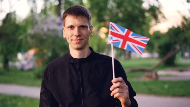 Retrato en cámara lenta del ciudadano orgulloso inglés feliz ondeando la bandera oficial del Reino Unido, mirando a la cámara y sonriendo. Concepto de personas y símbolos nacionales . — Vídeo de stock