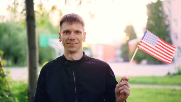 Retrato em câmera lenta do homem americano agitando a bandeira nacional dos EUA, sorrindo e olhando para a câmera. Conceito de jovens, cidadãos felizes, países e cidades . — Vídeo de Stock