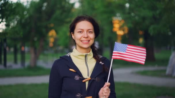 Retrato en cámara lenta de la orgullosa ciudadana estadounidense sonriente ondeando la bandera de Estados Unidos, mirando a la cámara y sonriendo. Árboles verdes, césped, senderos en el parque son visibles . — Vídeos de Stock