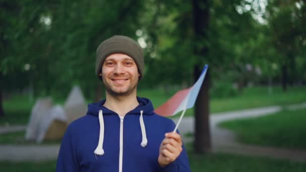 Slow motion portrait of handsome Frenchman waving official flag looking at camera and smiling with beautiful park in background. People, countries and nature concept. — Stock Video