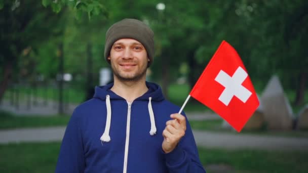 Retrato en cámara lenta del alegre viajero mundial con ropa casual sosteniendo la bandera suiza, mirando a la cámara con sonrisa alegre de pie en el parque de recreo . — Vídeo de stock