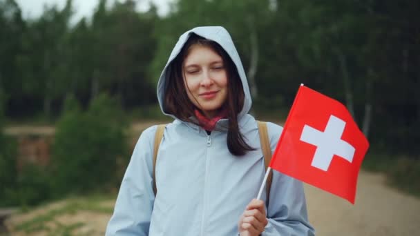 Retrato em câmera lenta de menina suíça bonita segurando bandeira da Suíça, sorrindo e olhando para a câmera. Bela paisagem com árvores verdes está no fundo . — Vídeo de Stock