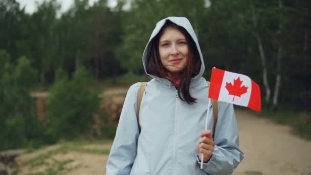 Retrato en cámara lenta de una chica bonita viajera sosteniendo la bandera canadiense, sonriendo y mirando a la cámara con hermoso paisaje natural en el fondo . — Vídeos de Stock