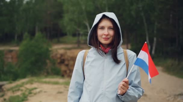 Retrato en cámara lenta de una joven atractiva sosteniendo la bandera de los Países Bajos, sonriendo y mirando a la cámara de pie sobre el fondo de la hermosa naturaleza . — Vídeos de Stock