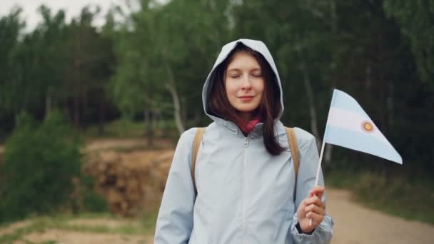 여성 세계 여행자는 바람에 비행 하는 아르헨티나 국기를 들고 웃 고 보고 카메라의 슬로우 모션 초상화. 아름 다운 숲은 백그라운드에서. — 비디오