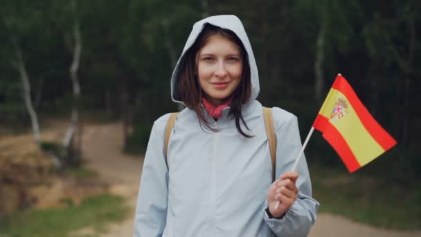 Slow motion porträtt av glada world traveller söt flicka i hooded coat viftande spanska flaggan och tittar på kamera med leende. Länder och resande koncept. — Stockvideo