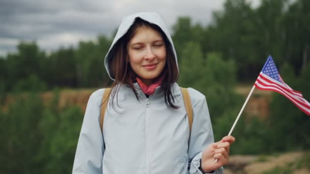 Retrato em câmera lenta de um cidadão americano orgulhoso agitando a bandeira dos EUA com um sorriso feliz e olhando para a câmera. Orgulho nacional, bela natureza, conceito de estilo de vida saudável . — Vídeo de Stock