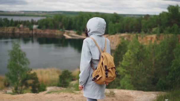 Slow motion porträtt av ung kvinna turist med ryggsäck till kanten av klippa med vacker utsikt över sjön och skogen. Resande, människor och natur koncept. — Stockvideo