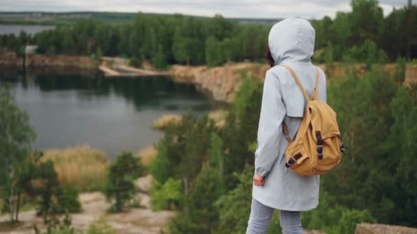 Przechylić w dół slowmotion portret szczupła dziewczyna, chodzenie na klifie z ją z powrotem do kamery i oglądania piękny krajobraz z jeziorem i drzew pod jej. — Wideo stockowe