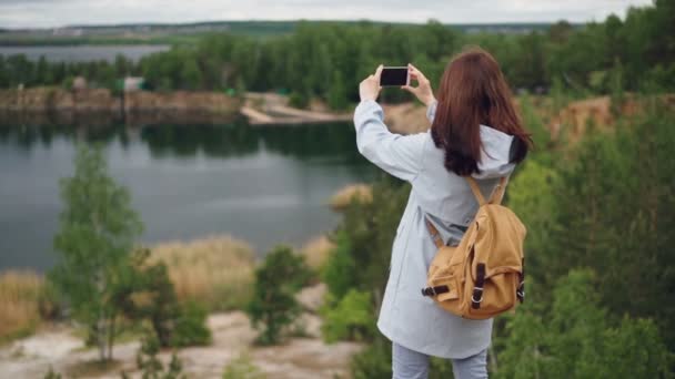 ナップザックのスマート フォンを使用して崖の上から美しい景色の写真を撮ると少女トラベラーのスローモーションの肖像画。美しい青い湖と緑の森が表示されます。. — ストック動画