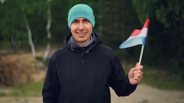 Retrato en cámara lenta del chico guapo en ropa deportiva ondeando la bandera de los Países Bajos, sonriendo y mirando a la cámara con hermosa naturaleza de primavera en el fondo . — Vídeos de Stock