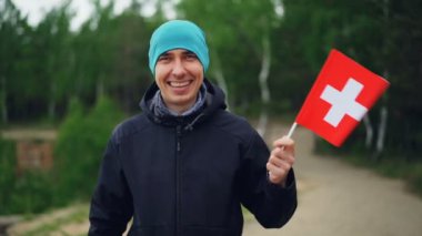 İsviçre bayrağı sallayarak ve kamera ayakta dışında yeşil ahşap yakınındaki bakarak gülümseyen Switzer ağır çekim portre. Gurur vatandaşlık, göç ve Vatandaşlık kavramı.