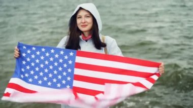 Amerika Birleşik Devletleri bayrağı tutarak ve dalgaları görünür ile deniz kıyısında ayakta hareket mutlu American Girl ağır çekim portre. Milliyet ve ülke kavramı.