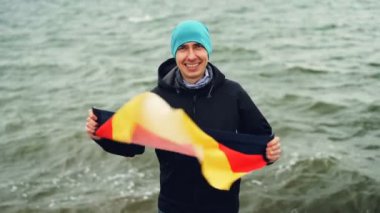 Ağır çekim portre Almanya bayrağı tutarak ve göl kenarında duran ve kameraya dalgalar arka plan üzerinde seyir sırasında gülümseyen neşeli Alman spor fan.