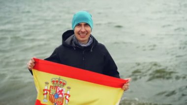 Ağır çekim portre mutlu İspanyolca fan holding deniz kıyısında duran ve gülümseyen İspanya bayrağı spor. Vatanseverlik, aktif yaşam tarzı ve insanlar kavramı.