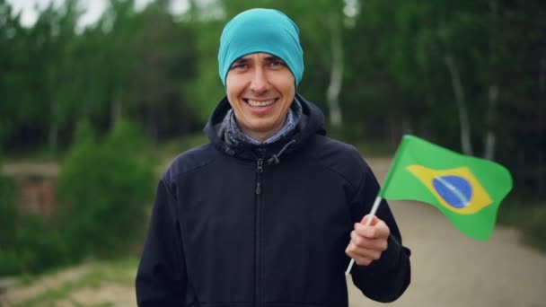 브라질 국기를 물결치 고 다행 미소와 함께 카메라를 보고 즐거운 사람 여행자의 슬로우 모션 초상화. 행복, 세계 각국 여행 방문 국가 개념. — 비디오