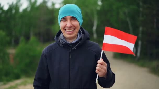 Ritratto al rallentatore di allegro turista in possesso di bandiera austriaca e guardando la fotocamera mentre in piedi vicino alla foresta verde in paese straniero e sorridente . — Video Stock