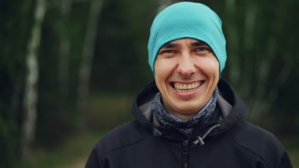 Primo piano ritratto al rallentatore di bel ragazzo in abiti casual cappello e giacca guardando la fotocamera e ridendo in piedi nella foresta. Concetto di uomo e natura . — Video Stock