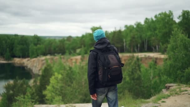 ナップザック坂道を歩き、崖山の湖と森の中でエキサイティングなビューを見ている若い男の観光客のスローモーションの肖像画. — ストック動画