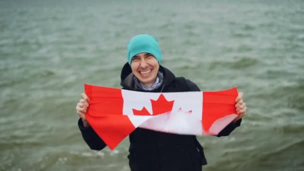 Retrato en cámara lenta de un joven feliz sosteniendo una gran bandera textil de Canadá, mirando a la cámara y sonriendo con olas de agua en el fondo. Concepto de turismo y naturaleza . — Vídeos de Stock